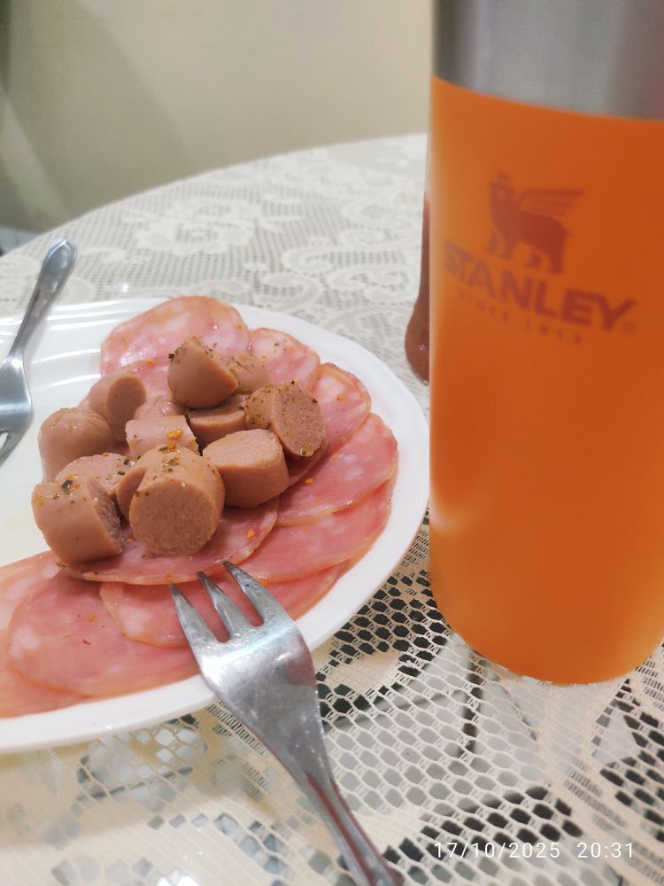 Pratinho branco com salaminho e rodelas de salsicha viena e copo Stanley laranja, em cima de mesa com toalha de renda cor palha.