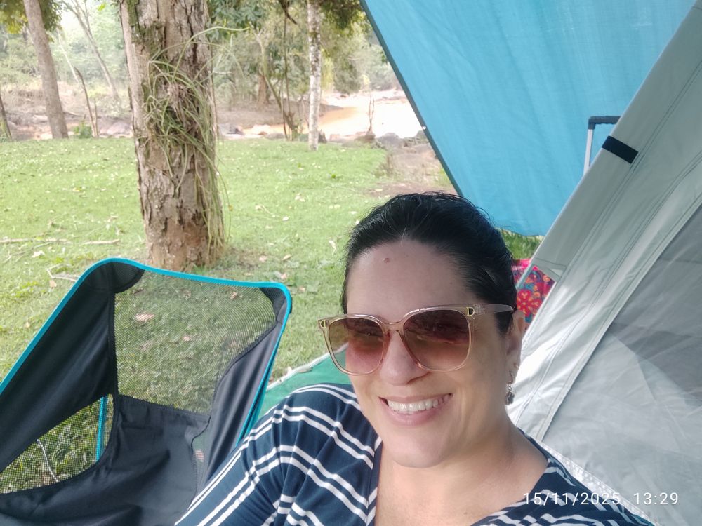 A imagem mostra uma mulher sorridente sentada em uma cadeira de camping em um ambiente externo natural. A mulher tem cabelos escuros presos, usa óculos de sol com armação marrom/rosada e uma camiseta listrada em azul e branco, está em primeiro plano, sorrindo para a câmera. Ela está sentada em uma cadeira de camping preta e azul. Ao fundo, há um cenário de camping com árvores, grama e um rio. Parte de uma barraca e de uma uma lona azul é visível no lado direito da imagem.