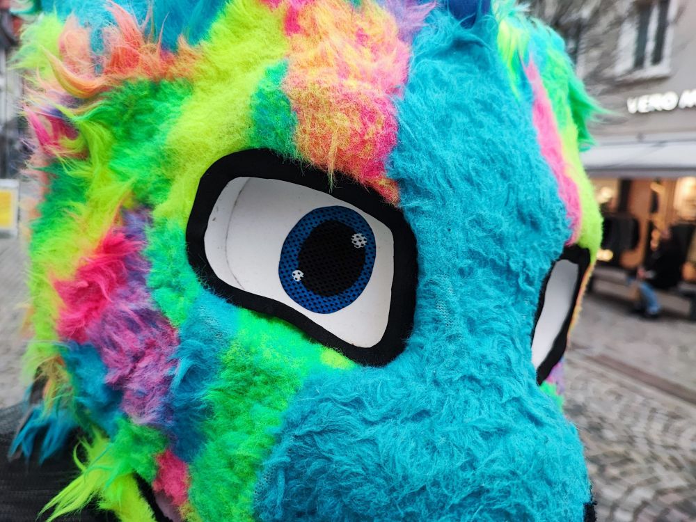 A fursuiter with a rainbow-colored dragon suit looks into the camera. The caption reads: I have a keen eye on you 😊😁