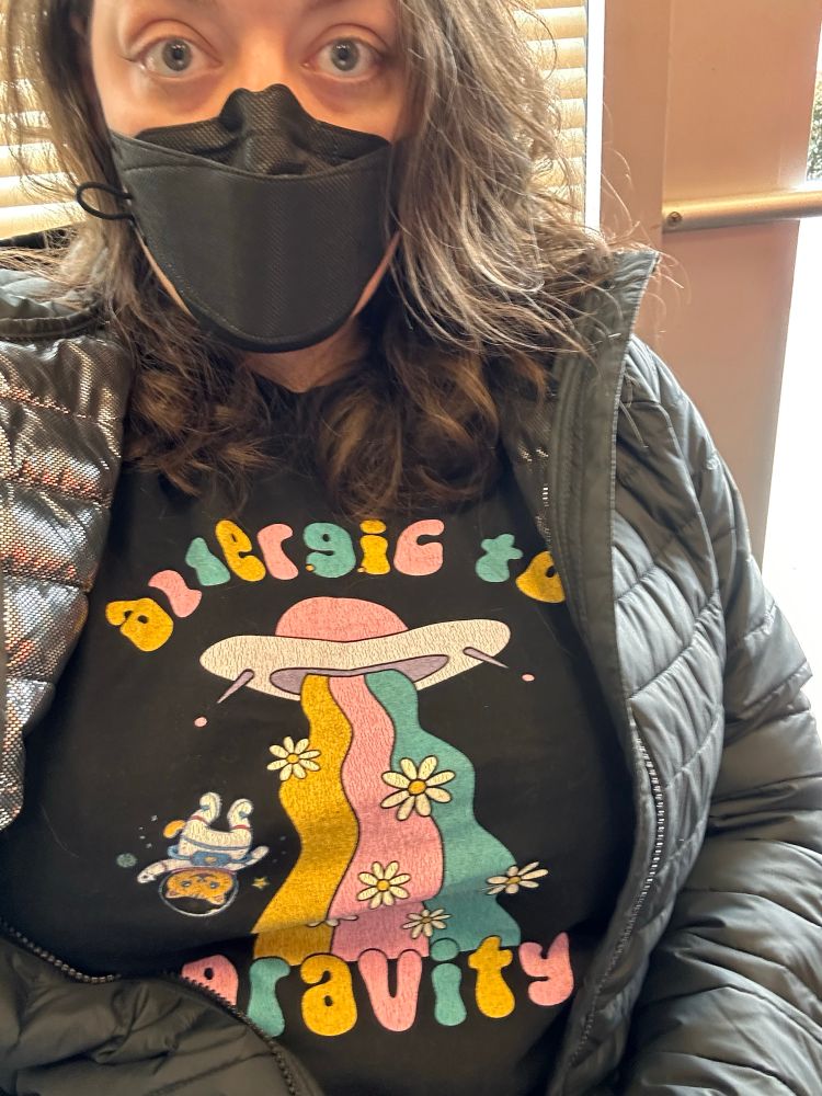 A white woman wearing a mask and winter coat over a tshirt with a spaceship and a floating cat astronaut that says "allergic to gravity"