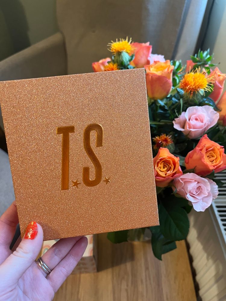 A hand holds an Orange sparkly box with letters TS on front with a bouquet orange and pink roses in the background 