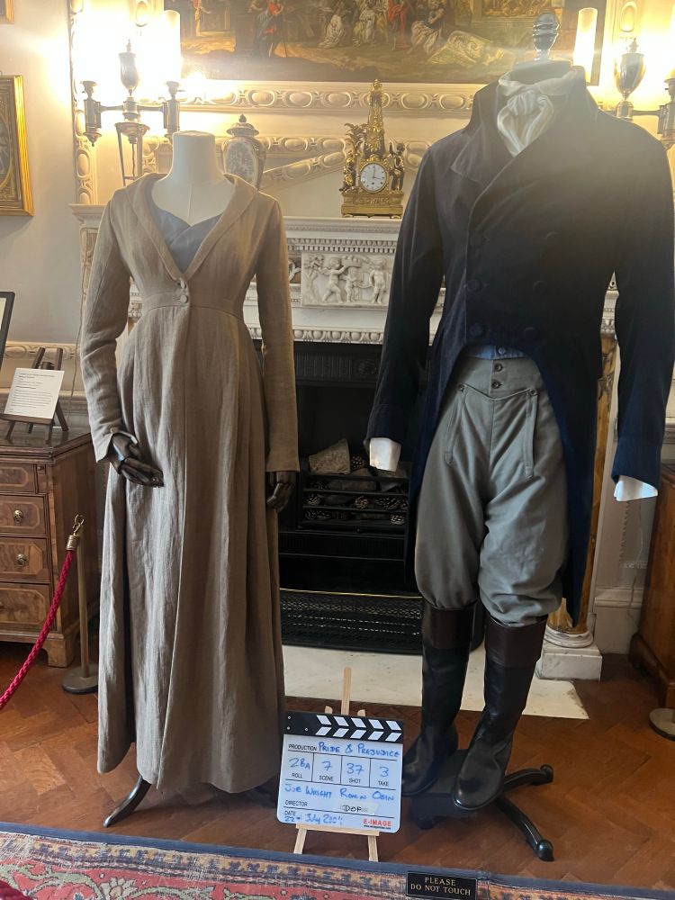 On the left the photo shows a copy of the outfit worn by Keira Knightley playing Elizabeth Bennet in the 2005 Pride and Prejudice film. The outfit is mainly a tawny coloured long coat. There is a hint of a greyish/blue dress underneath. 
On the right is the actual outfit worn by Matthew Macfadyen when he played Mr Darcy. It shows a whitish grey cravat, dark blue/black tailcoat, greyish breeches and black boots. There is a hint of a light blue waistcoat underneath.
Beneath both outfits sits a clapperboard saying Pride and Prejudice, roll 28A, scene 7, shot 37, take 3, Joe Wright, Roman Osin, 22 July 2004. 
The costumes are from the scene is when Lizzie and Darcy argue after the Rosings church service (when Colonel Fitzwilliam unknowingly indicated Darcy separated Bingley from Jane). The scene was filmed at the Temple of Apollo at Stourhead. 