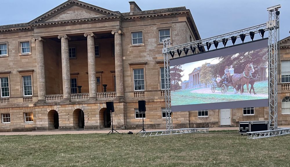 Basildon Park building shows on left, with its four large pillars and first floor entrance. On the right is an outdoor screening of the 2005 Pride and Prejudice, taking place on the grass opposite the house. The scene shows a horse and carriage departing “Netherfield” (Basildon Park is used as the location). 
