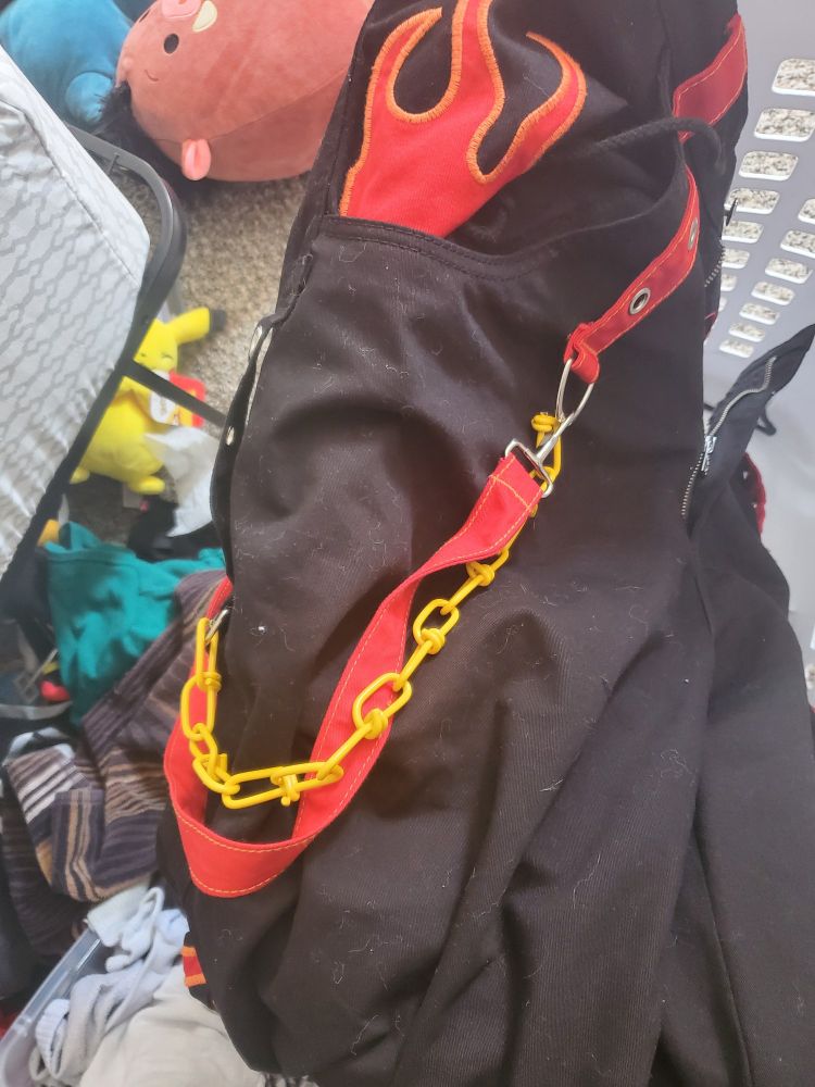 A black pair of pants with flames on the pockets. There is a red strap from the front of the pants to the back, and a yellow chain from the front to the side.