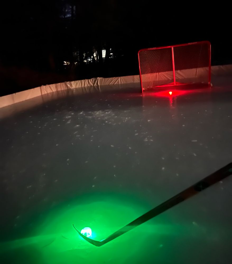 A backyard hockey rink at night. In the foreground a hockey stick cradles a puck that is glowing bright green. In the background a bright red glowing pucks rests in a hockey net.