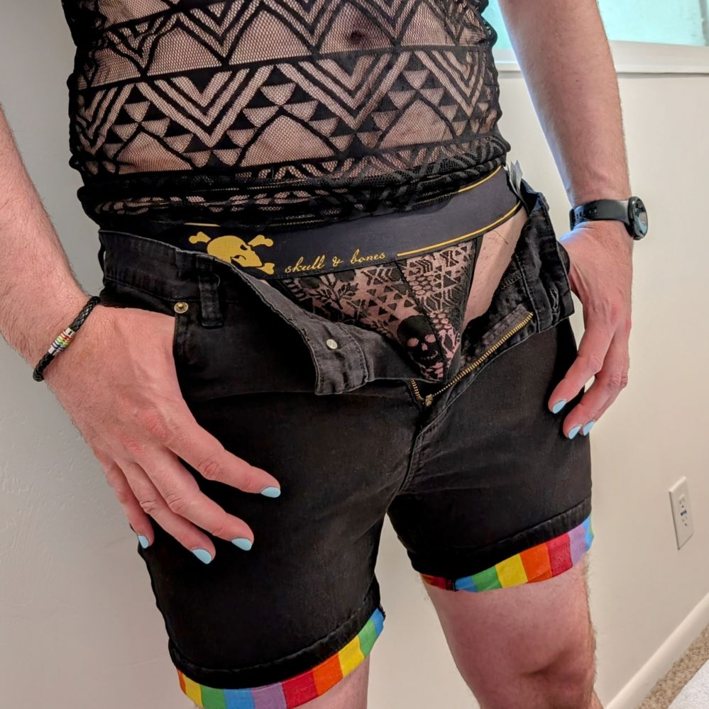 A picture of a man's lower torso and thighs. He is wearing a black, lacey top and black shorts with the hems turned up to reveal rainbow stripes. The fly is wide open revealing a black lace Skull and Bones thong with his semi erect cock visible through the lace.