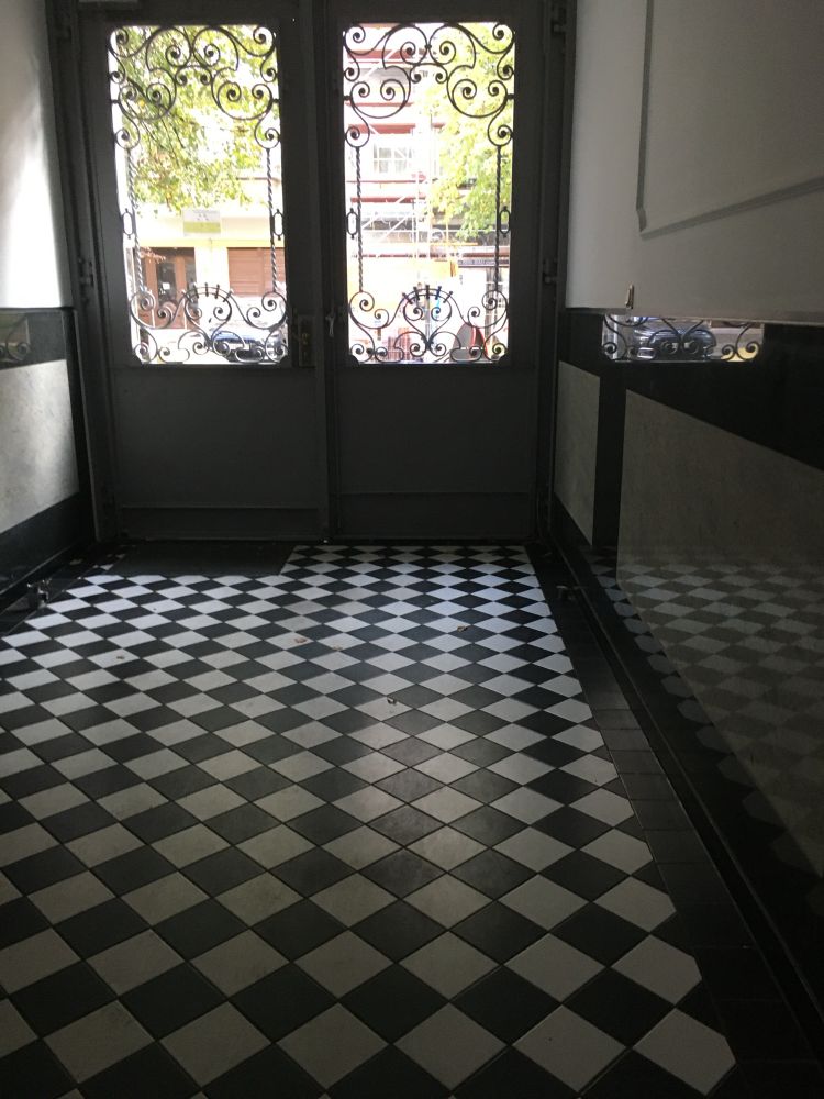 Das Foto zeigt den Eingangsbereich eines Gebäudes. Der Boden ist mit einem schwarz-weißen Schachbrettmuster gefliest. Rechts und links sind dunkle Wandverkleidungen mit hellen Paneelen. Am Ende des Flurs befinden sich zwei verglaste Türen mit dekorativen, geschwungenen Metallgittern. Durch die Türen sieht man Bäume und ein Gebäude draußen bei Tageslicht.