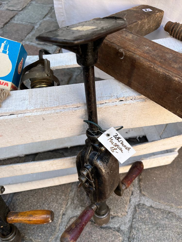 Auf dem Bild liegt ein altes mechanisches Werkzeug auf einer weißen Holzkiste, vermutlich ein handbetriebener Bohrer oder Schraubstockaufsatz mit zwei Holzgriffen und einem Metallgestänge. Daran hängt ein kleines Etikett mit handgeschriebener Beschriftung. Im Hintergrund sieht man weitere alte Werkzeuge und Kopfsteinpflaster.
Gesehen auf der Potsdamer Antikmeile.