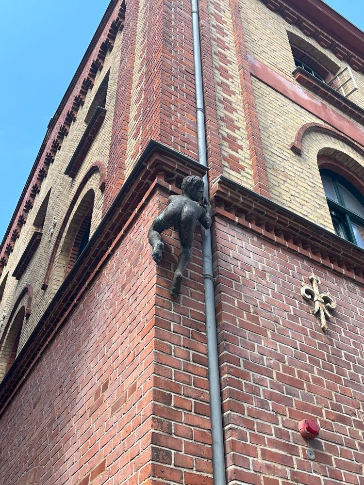 Auf dem Foto ist die Ecke eines Backsteingebäudes zu sehen. Das Gebäude besteht aus rotbraunen und beigefarbenen Ziegeln und zeigt eine historische oder industriell anmutende Architektur mit Rundbogenfenstern und Ziermauerwerk.

An der Gebäudeecke ist eine Regenrinne angebracht, an der eine Skulptur befestigt ist: Sie stellt eine menschliche Figur dar, die so wirkt, als würde sie an der Regenrinne hinaufklettern. Die Figur scheint aus Metall oder Bronze zu bestehen.

Rechts an der Wand ist außerdem ein goldenes Zierelement in Form einer stilisierten Lilie (Fleur-de-Lis) angebracht, und weiter unten befindet sich ein roter Alarmknopf oder Sensor. Der Himmel ist klar und blau.