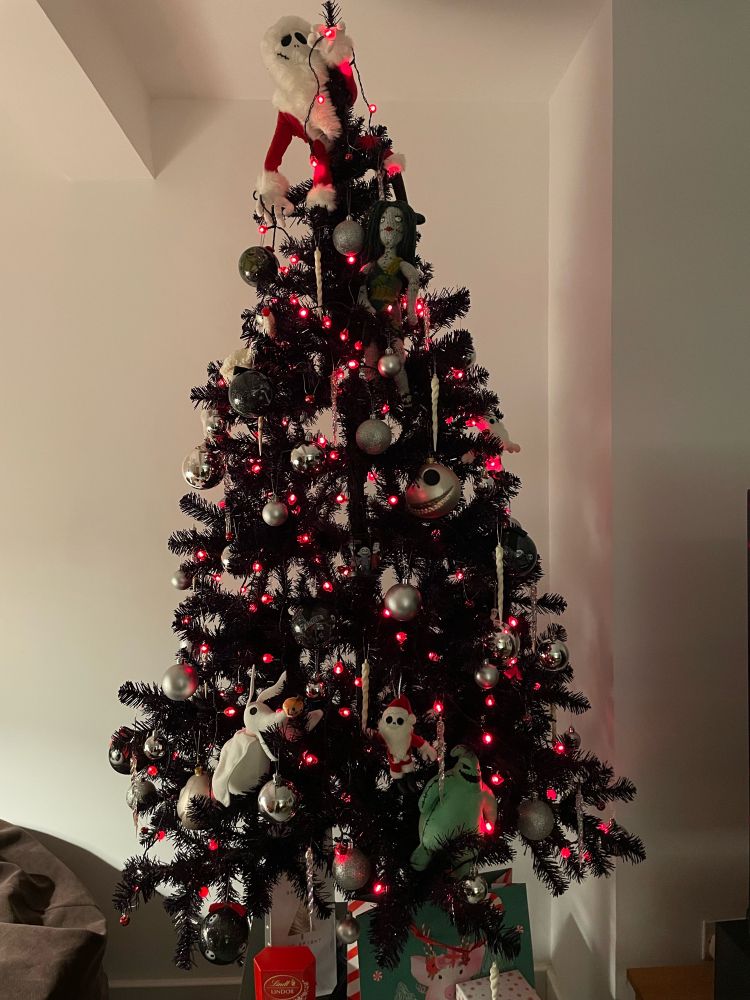 An artificial Christmas tree (looks black but it’s actually a very dark purple) with red lights, silver baubles, and decorations themed around the Tim Burton animated movie The Nightmare Before Christmas. A Jack Skellington plushie climbs to the top of the tree holding a string of lights, and a hand crocheted Sally sits beneath him. At the bottom of the tree, the ghost dog Zero keeps an eye on mischievous Mr Oogie Boogie.