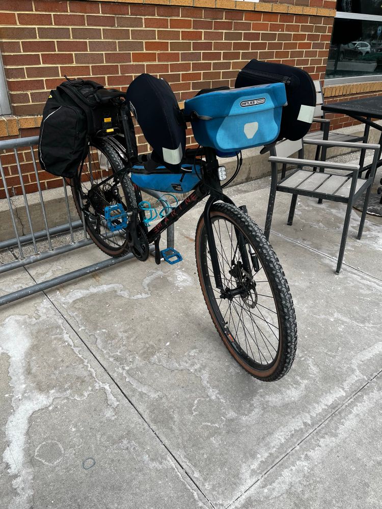 A black drop bar gravel bike with black bar end mitts and panniers, and a blue handle bar bag and frame bag, with the back wheel in a bike rack in front of a brick store. 
