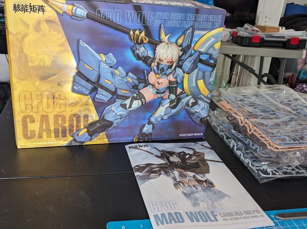  box, instructions, and still-bagged sprues rest on a black work surface. The instructions have the text Mad Wolf in blue on the bottom, while the box shows a picture of a scantily-clad armored girl with wolf ears lunging forward, an axe resting on her shoulder, against a blue background, while a band of yellow is on the left side of the box, and bears the words CF06 Carol