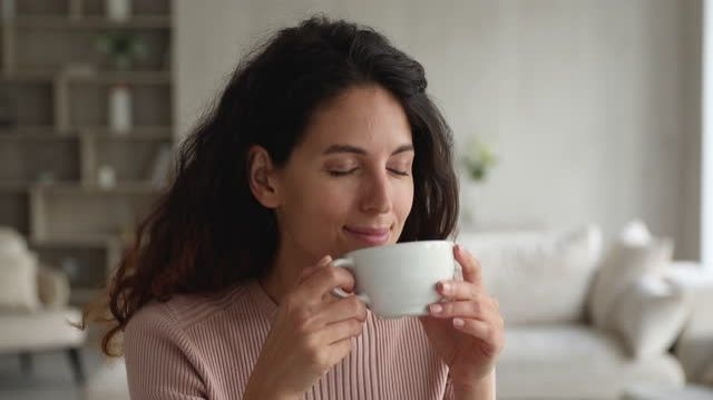 Eine junge Frau mit schulterlangen dunkelbraunen Haaren, die einen dezent rosafarbenden Pullover trägt und im Wohnzimmer mit geschlossenen Augen voller Genuss ihren Tee trinkt
