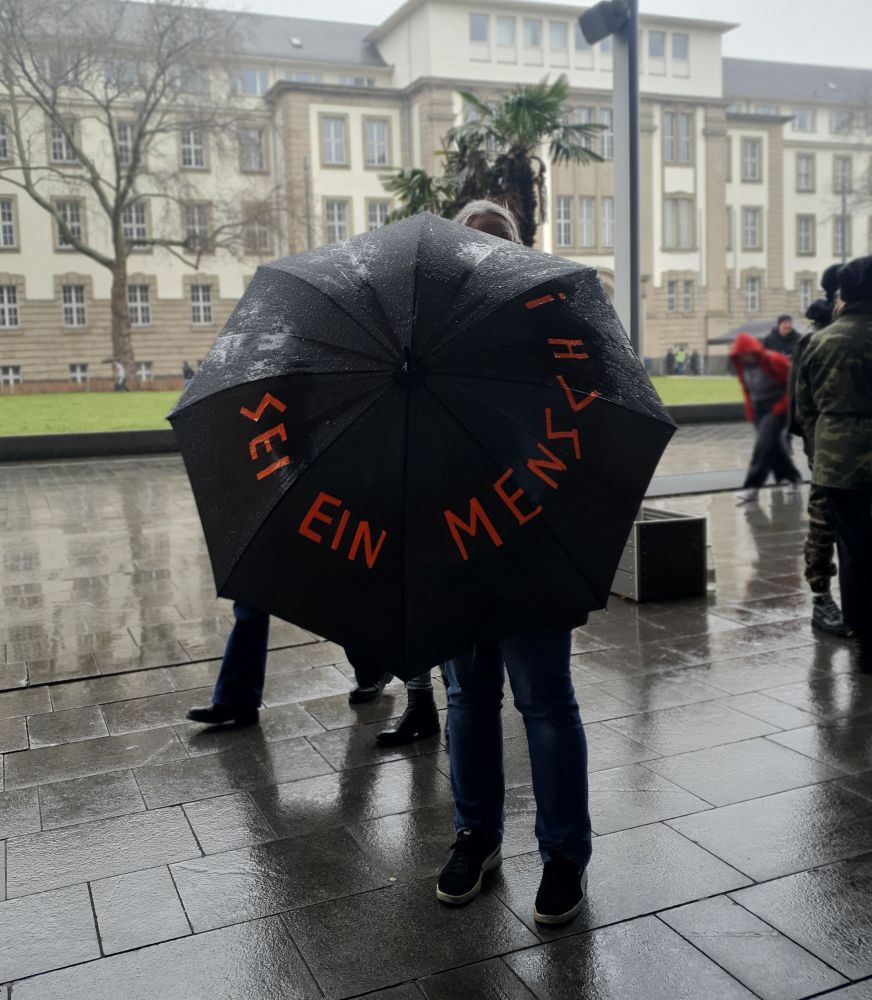 Eine Person hinter einem ausgespannten, schwarzen Regenschirm, auf dem mit orange 'Sei ein Mensch!' steht.