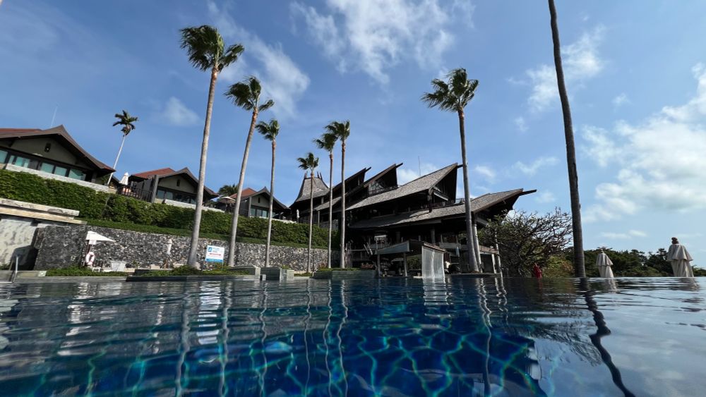 An infinity pool looking up at the resort. Private suites sit above the pool, also with their own private pools. A large structure 3-4 stories tall in the background climbs up the steep hillside. The structure is in the style of a tropical Thai building with sloping tiered roofs for each level. Palm trees scattered on the property. 
