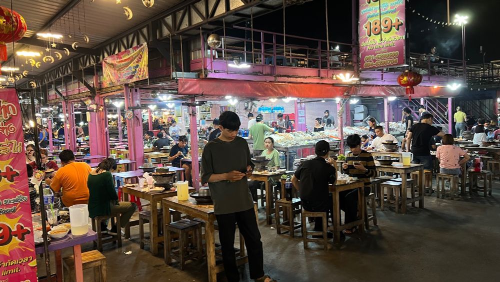 Tables surrounding a hotpot restaurant at the night market. Inside the restaurant, tables of ingredients are laid out for patrons to pick and choose for their hotpots which they cook at the table. 