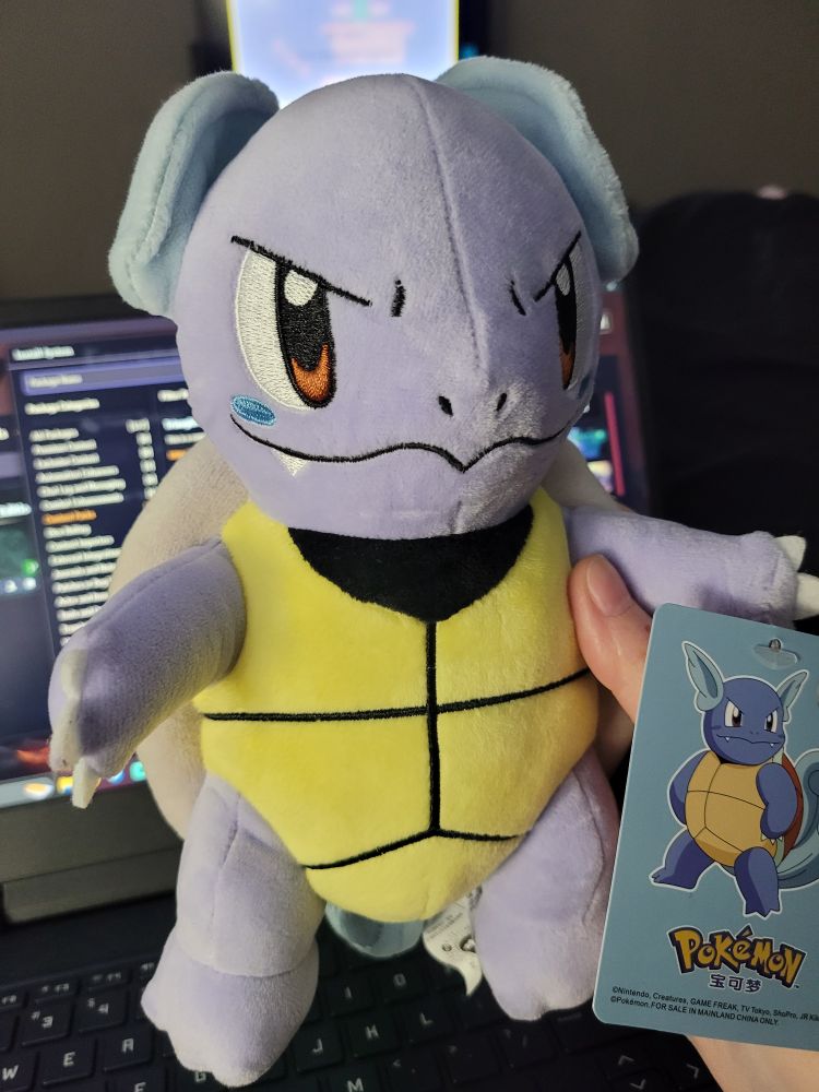 A photo of my hand holding a wartortle plushie over my open laptop with the Foundry (a virtual TTRPG program) on the screen. You can see my TV screen on in the background, but I have no idea what's on it. Knowing me, it is YouTube and could be anything from Sheep farming videos to Watts The Safeword.