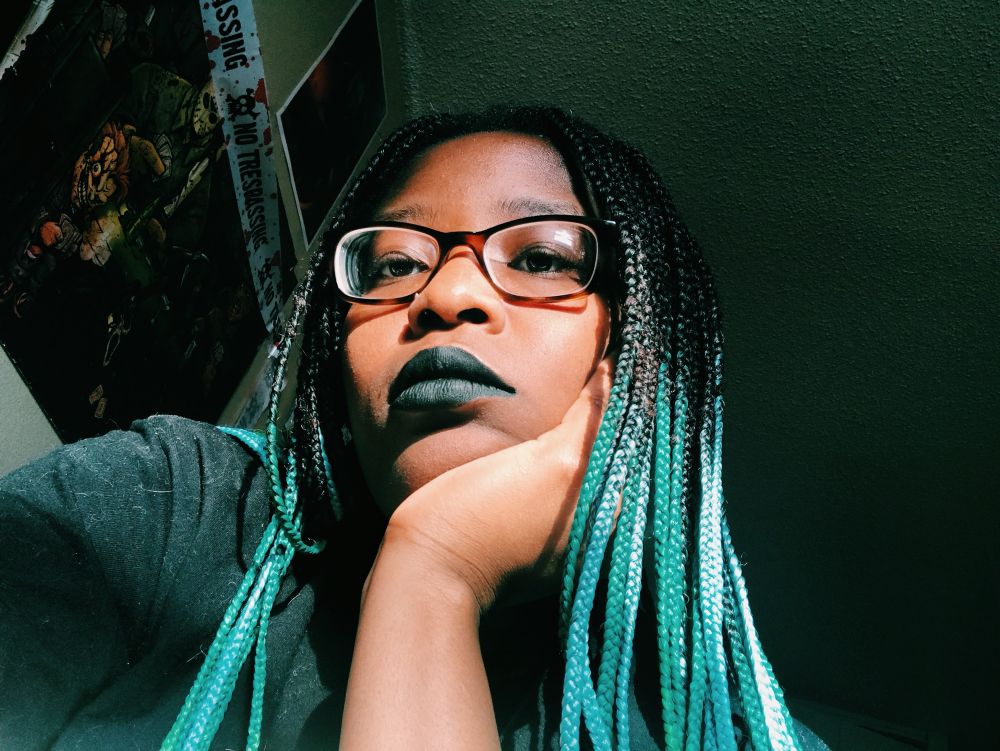 selfie of me, with tortoise shell glasses, black/blue ombré braids, black lipstick, one nose ring, sitting in front of horror movie posters. this is from like 2019 I think