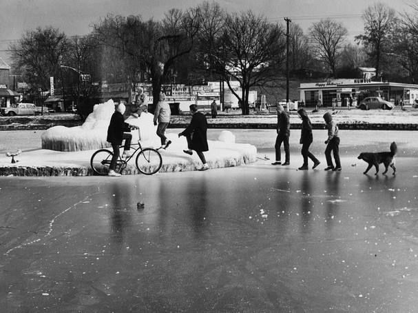 A black-and-white photograph shows a group of people on a frozen pond or lake. One person is riding a bicycle across the ice while others walk, balancing on the slick surface. Some individuals appear to be cautiously stepping onto or off a raised ice platform in the center of the image, which resembles a fountain or frozen structure. A dog is walking nearby on the ice, following the group. In the background, leafless trees and small commercial buildings, including a store with a visible sign, indicate a winter setting in a town or suburban area. The frozen pond reflects the figures, adding depth to the scene.
