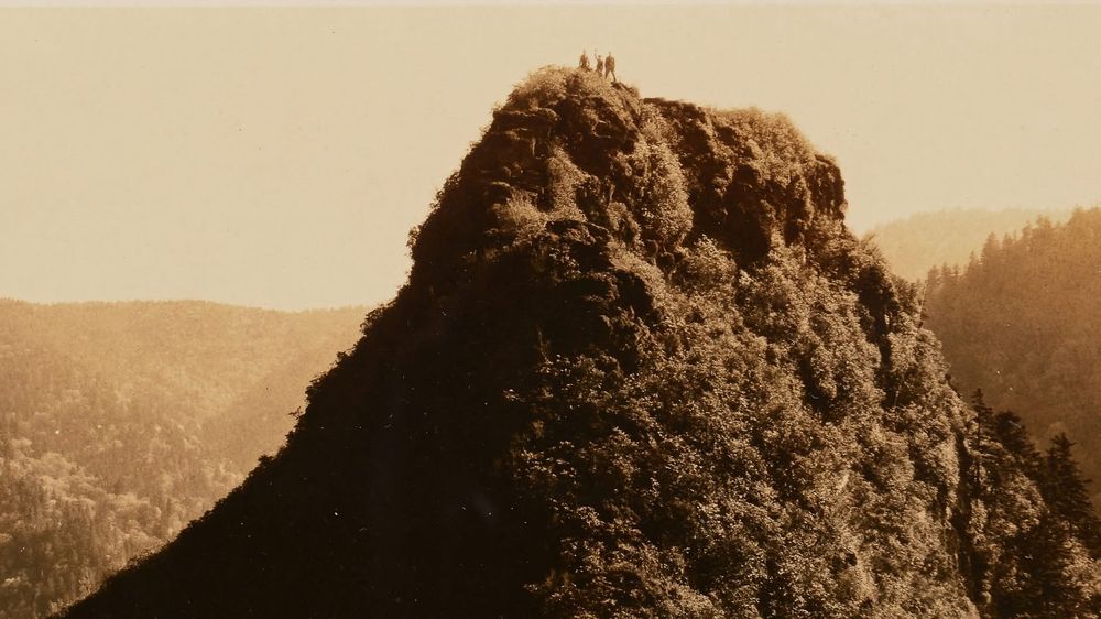 This is a sepia-toned photograph depicting a steep, rocky peak covered with foliage. At the top of the peak, a few individuals are visible, silhouetted against the sky, giving a sense of scale and adventure. The surrounding area is filled with hilly, forested terrain, creating a majestic and serene landscape.