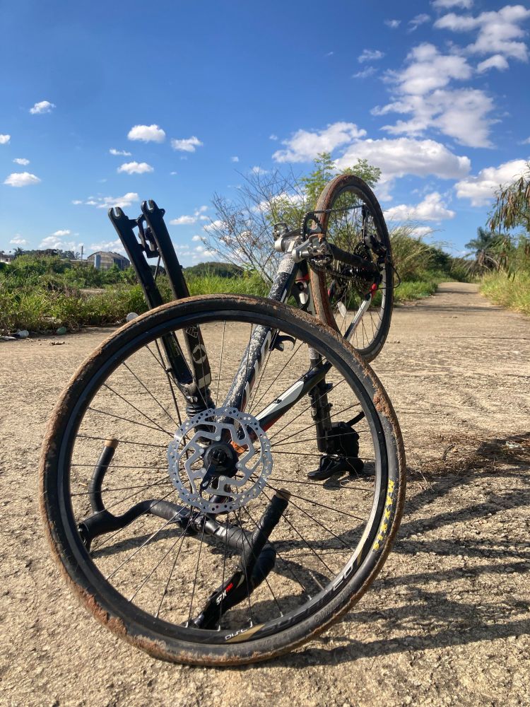 Fotografia colorida de uma bicicleta de ponta cabeça, como guidão e o selim apoiados no chão. A roda dianteira está desmontada e apoiada no garfo. Ao fundo, um céu azul e vegetação. 