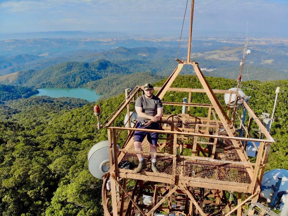Fotografía aérea de um homem branco no topo de uma torre de telecomunicações no Pico do Gil, em Guarulhos.
A foto foi feita numa manhã ensolarada e mostra, em primeiro plano, o topo da torre com o homem okhanpara a câmera. Em segundo plano, a mata nativa da Serra do Itaberaba e o lago da Ambev. 