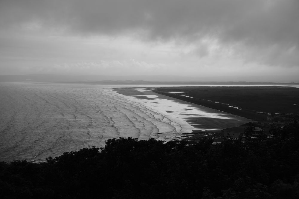 Black and white image taken from the cliff above a large beach. Lots of waves are rippling in. In the distance storm clouds are brewing 