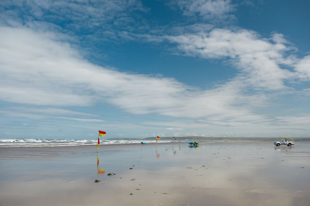 a wide open beach with red and yellow safety flags, an RNLI coastguard truck and groups of trainee surfers walking down the beach
