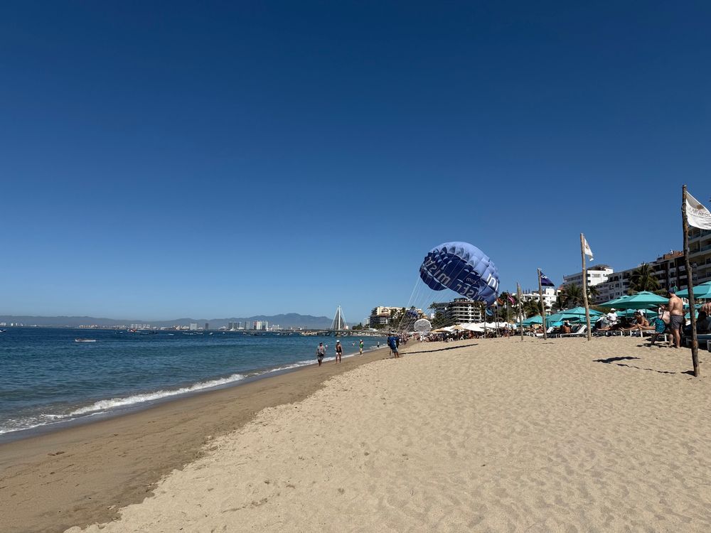 Beach photo looking along the waterline. The ocean is on the left a d there are various building and beach umbrellas nestled along the right. 
A parasailing vendor can be seen walking his parachute, inflated by the breeze, towards the camera. 