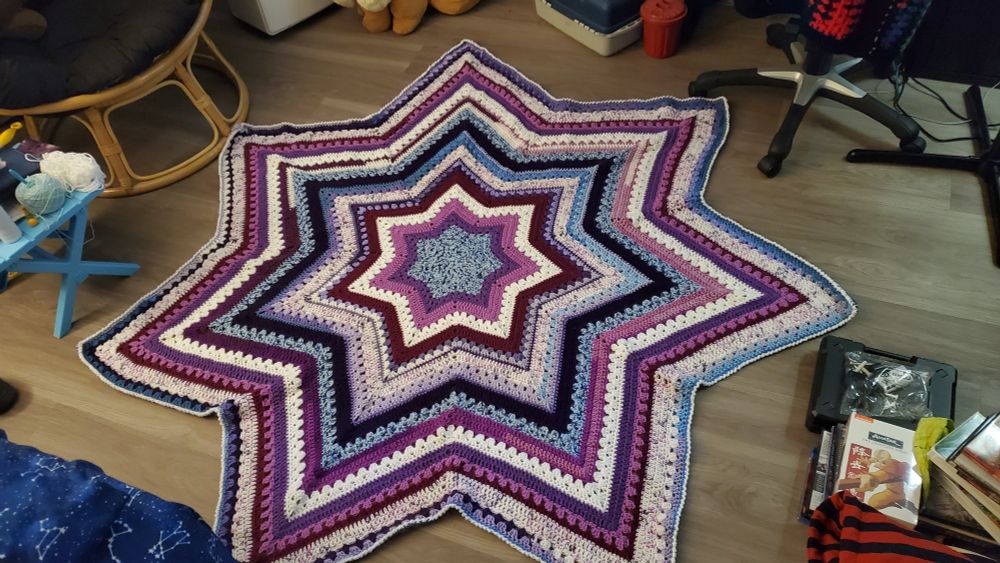 A large, 7-pointed star blanket sitting on a vinyl flooring. The blanket is comprised of many purples, blue, and white. 