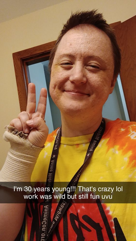 It's a selfie of Phoenix. He is wearing a red, orange and yellow tie dye shirt (missing the green or it'd be rainbow lol) and he is smiling and throwing up a peace sign. He has a black and white key lanyard from an ancient Animecon Minneapolis convention around his neck. His wrist compression sleeve is barely holding on by a thread. 