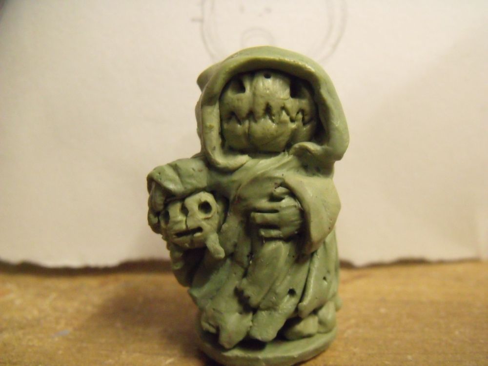 A green stuff sculpt of a pumpkin person wearing a cloak and holding a pumpkin with legs under one arm.