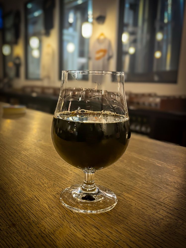 Deep dark imperial stout in a curved beer glass on a deep brown wooden counter.
