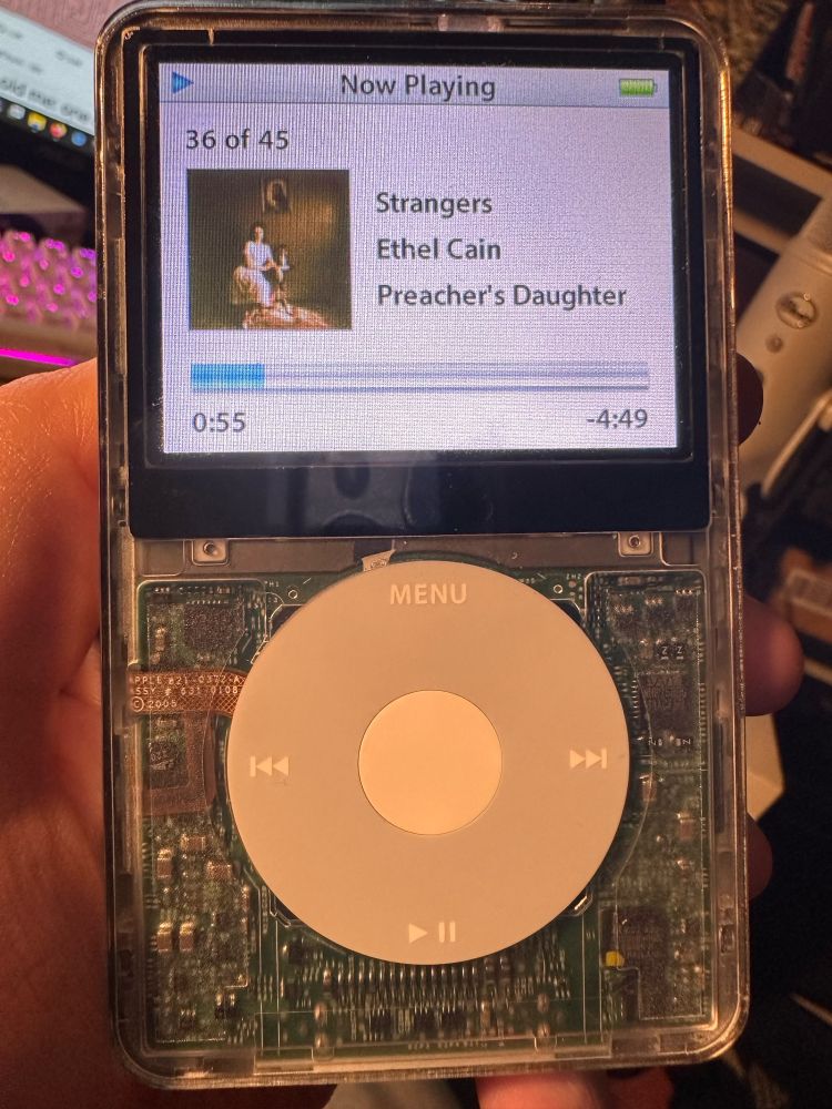 front view of my ipod classic, clear case showing the circuitry inside, screen on and showing the song Strangers by Ethel Cain playing
