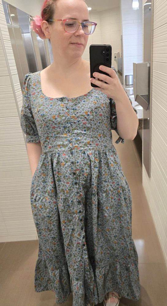 A female presenting white human stands in a bathroom mirror, looking at their phone. They are wearing a blue dress with green buttons down the front. It has a scooped neck, puffy sleeves and a tiered skirt.