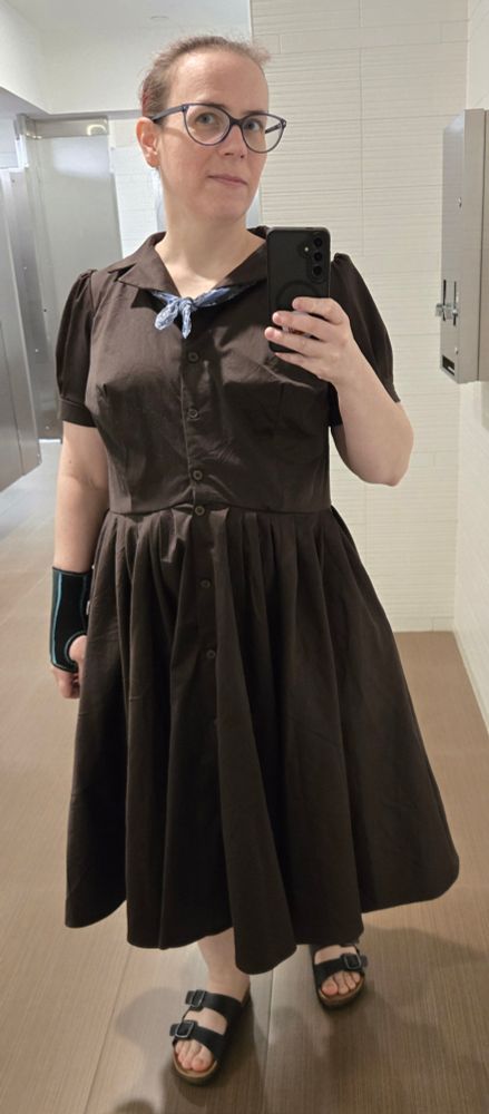 A white female-presenting person stands in a bathroom mirror dressed in a dark brown shirt dress with a very full skirt, with a blue scarf tied at the collar. They are also wearing sandals and a wrist brace.