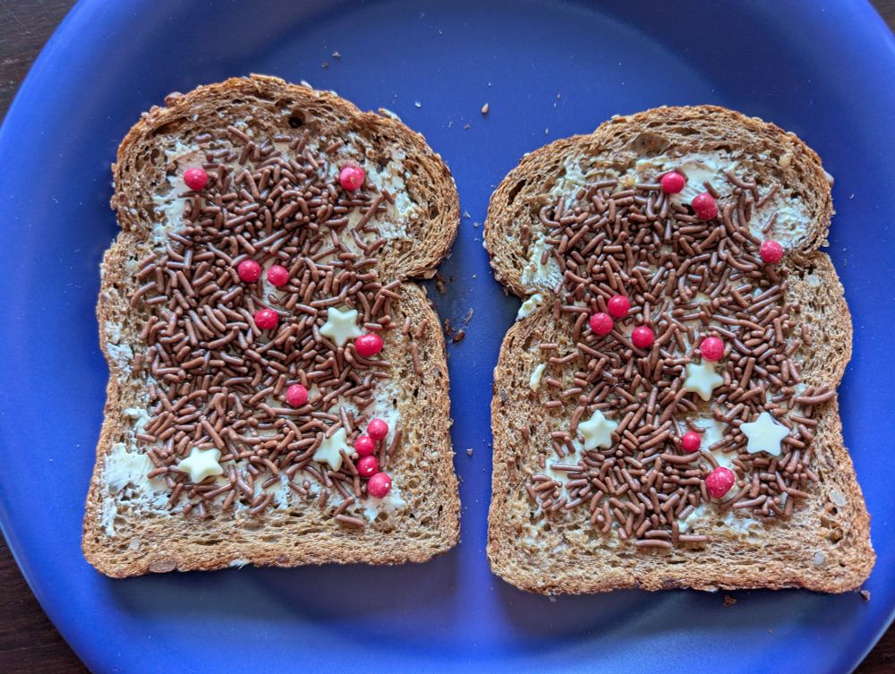 Twee multivolkoren sneetjes brood op een blauw bord, waarop hagelslag ligt met rode bolletjes en witte sterretjes van chocolade.