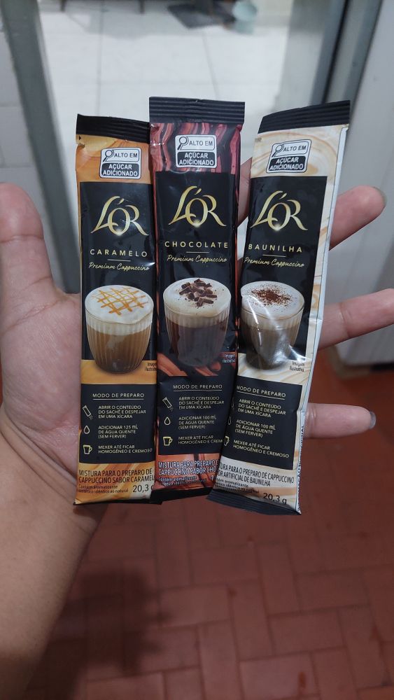 uma mão segurando três embalagens de formato retangular de capuccinos da marca Lor, sendo o primeiro de caramelo, o segundo de chocolate e o terceiro de baunilha