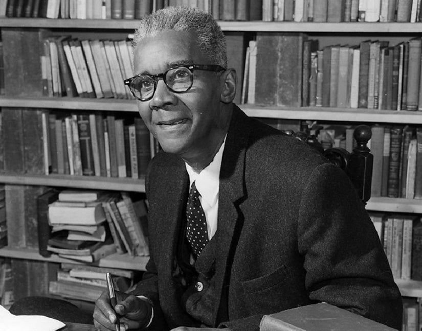 A Black man (C. L. R. James) with glasses and in a suit, with a wall of books on the background. 