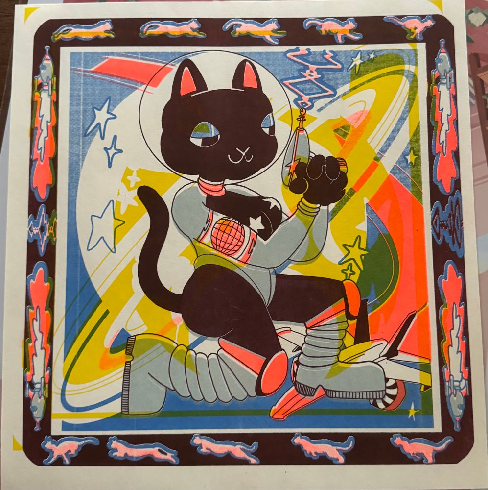 Risograph print in blue, light blue, black, neon peach, and yellow. A black cat in a sexy spacesuit and big boots. She has a ray gun. There are stars and planets in the background. The tborder is black; the top and bottom have pink cats in various stages of leaping, and the sides have rocket ships.