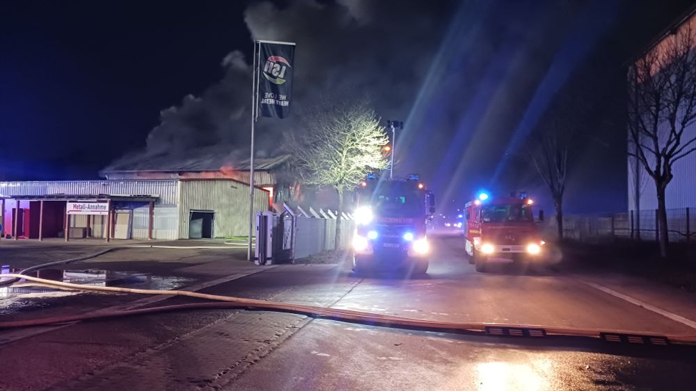 2 Feuerwehrfahrzeuge mit Blaulicht im Einsatz, brennende Industriehalle im Hintergrund