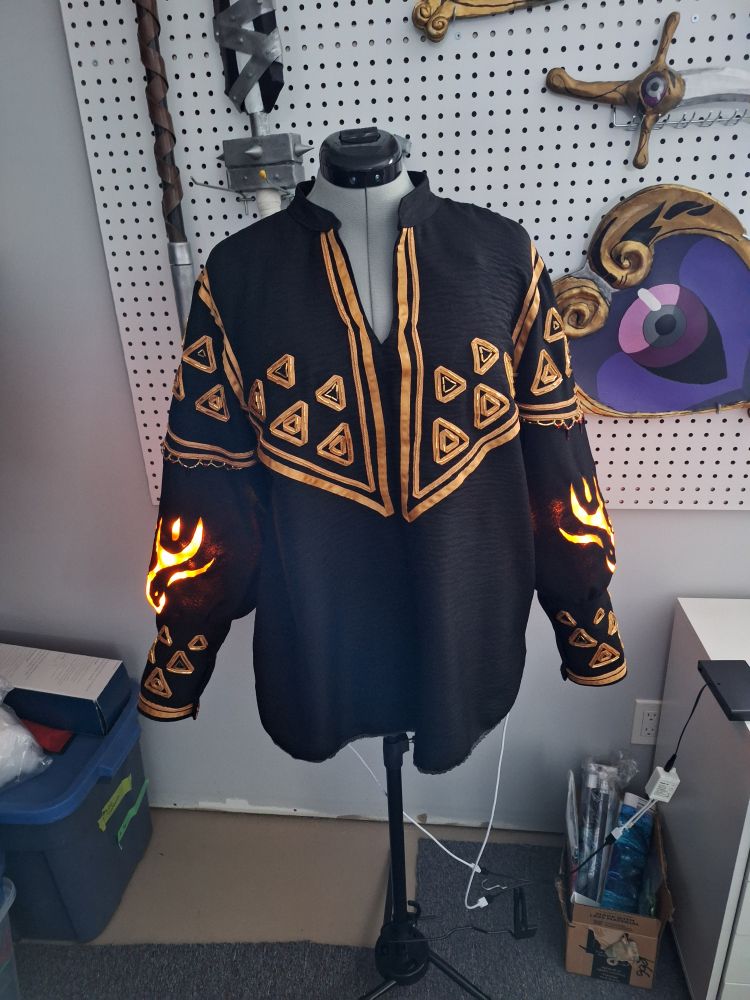 Shirt hanging on a dress form. It is based on Link's Ember Armour shirt from Legend of Zelda.
The shirt is a black tunic with a short stand collar and poofy sleeves that have somewhat long cuffs. There are gold ribbons used as borders and triangles decorating the whole shirt. The sleeves have flame cutouts that are lit by LEDs to make them glow.
There is beading on top of all of the triangles and along the sleeves