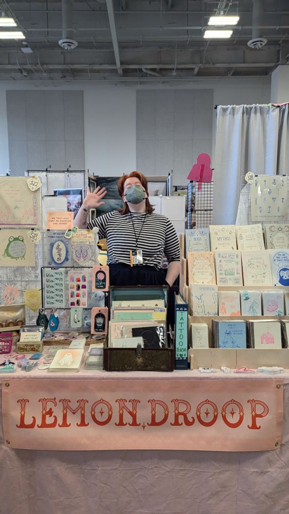 Lemon in a striped shirt waving from behind their artist alley table with a big sign that says lemon.droop