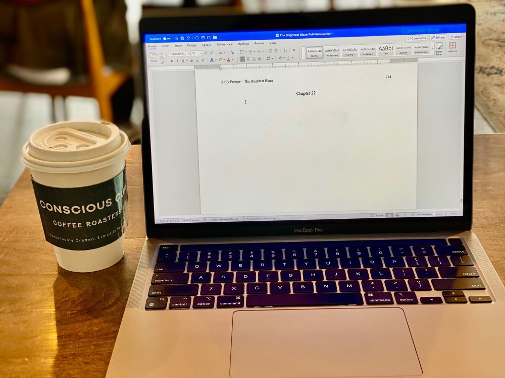 Photo at a coffeehouse of an open laptop with a Word document on the screen with "Chapter 22" typed on it. A to-go cup with a Conscious Cup sleeve around it is sitting beside the laptop.
