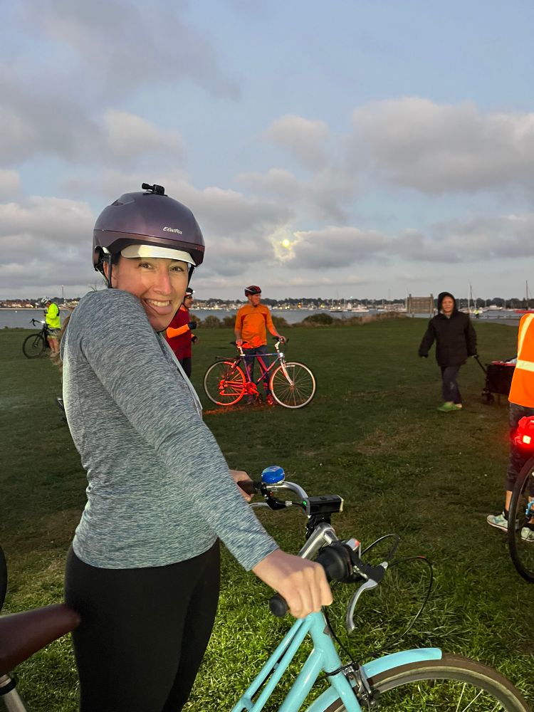Emily stands sideways to the camera, holding her bike handlebars, grinning, with the full harvest moon in a cloudy sky behind her. She is in a field with other bikers behind her. 