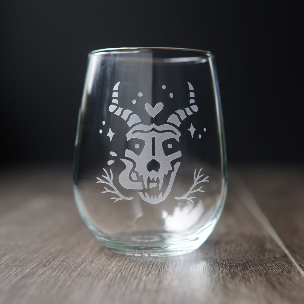 A skull with twisted antlers and long fangs engraved into a stemless wine glass. The skull has a long tongue with saliva spraying off it, and a cute little heart above its head. 