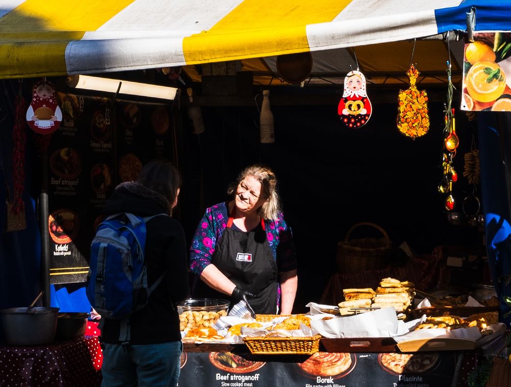 Image of a stall holder at Cambridge Market who is illuminated by the afternoon sun in conversation with a customer 