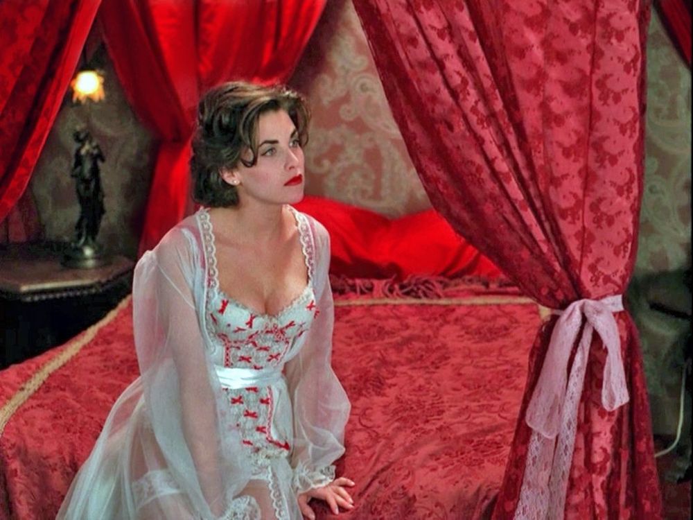 A still from Twin Peaks featuring the character Audrey Horne, played by Sherilynn Fenn, on a fancy silk bed, wearing white lingerie with sheer sleeves and a red bow pattern