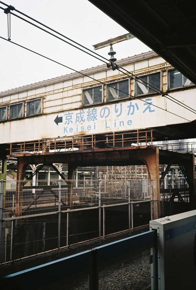 A bridge going over railway tracks. Lot of rusty metal. The bridge is a covered walkway. Underneath some Japanese characters, text in blue says 'Keisel Line'.