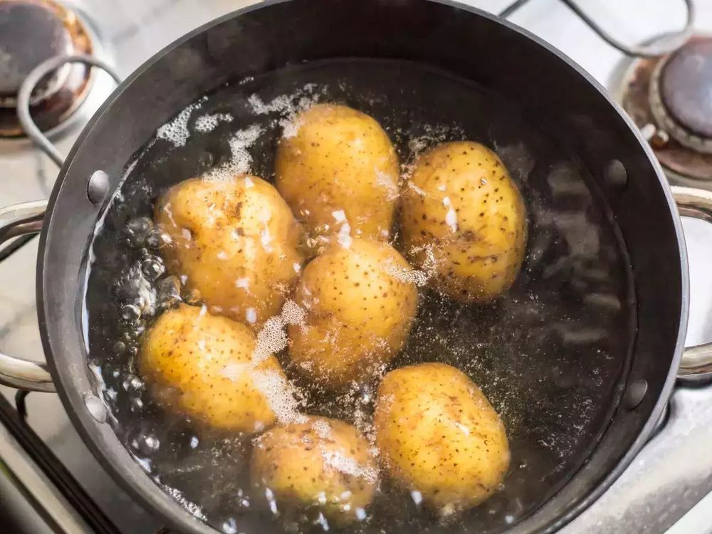 WAPs, or wet-ass potatoes. Seven small, golden-colored spuds (perhaps Kennebecs, News, or Yukon Golds). They’re submerged in a pot of boiling water on top of an old white stove with what look to be gas burners.